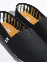 Alpargata de lona negra con suela de goma blanca flexible. Presenta una etiqueta pequeña con el logo de la marca en el lateral y plantilla interior de símil cuero.