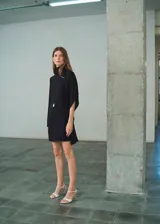 Vestido corto negro de corte recto con cuello alto y mangas asimétricas. Presenta cierre posterior con cremallera y botones.