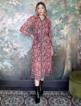 Vestido camisero midi de gasa estampada con flores rojas y blancas sobre fondo marrón. Tiene cuello con solapa, abertura central con botones, mangas largas y lazo de ajuste en la cintura.