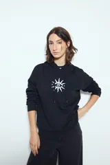 Canguro negro de calce relajado con capucha y cordones ajustables. Presenta un estampado gráfico frontal de un ojo con rayos de sol y la frase "STAY WEIRD".