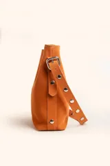 Cartera tipo balde color tostado, confeccionada en gamuza, con correa ajustable decorada con tachas metálicas plateadas.
