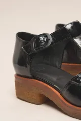 Zapato tipo guillermina de cuero charol color marrón oscuro, con tira T y hebilla ajustable en el lateral. Posee puntera cuadrada y suela de goma color caramelo con taco bajo.