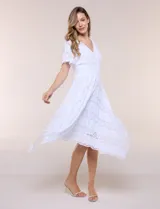 Vestido midi blanco de estilo romántico confeccionado en poliéster con elastano, con detalles de encaje floral. Cuenta con escote cruzado en V, mangas cortas acampanadas, cintura elastizada y falda evasé con terminación asimétrica en picos.