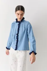 Camisa de jean azul claro con detalles de crochet en color azul oscuro en el cuello, la botonera frontal y los puños. Presenta un corte recto, mangas largas y botones de madera.