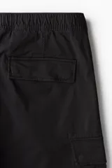 Pantalón cargo negro de corte slim fit, con cintura elástica y cordón ajustable. Presenta bolsillos al bies, bolsillos cargo en las piernas con solapa y botones a presión ocultos, y bolsillos traseros con solapa. Las piernas son ajustadas con costuras y pinzas en las rodillas.