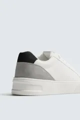 Zapatilla deportiva blanca con suela de plataforma gruesa y texturizada. Presenta detalles en gamuza gris en el lateral y un parche negro en el talón.