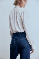 Pantalón de jean estilo wide leg con tiro alto, confeccionado en denim de algodón rígido. Presenta bolsillos delanteros con detalle de ribete trenzado, pliegues frontales y traseros, y cierre con doble botón.