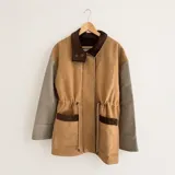 Chaqueta de silueta relajada en combinación de paño de lana marrón y pana de algodón en color marrón oscuro, con mangas grises. Cuenta con cuello alto, bolsillos frontales amplios y cordón regulable en la cintura.