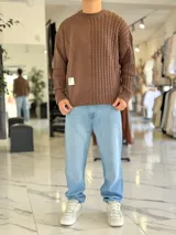 Sweater de punto oversize color marrón, con cuello redondo y diseño combinado: la mitad derecha cuenta con tejido de ochos y la izquierda es lisa. Incluye una etiqueta decorativa blanca en el frente inferior derecho.
