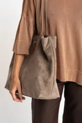 Bolso tipo tote de gran tamaño en color camel, confeccionado en cuero vegano con acabado efecto gamuza. Presenta doble asa larga para llevar al hombro y una placa metálica pequeña con el logo de la marca en el frente.