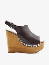 Sandalias de cuña alta de madera, color marrón, con capellada de cuero, tira trasera ajustable con hebilla y detalles de tachas metálicas plateadas en el contorno de la suela.