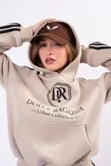 Canguro oversize color beige con capucha, bolsillo frontal tipo canguro y estampado gráfico en el pecho con el logo de la marca. Presenta tres rayas negras decorativas en las mangas.