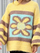 Buzo de tejido de punto con textura peluda, de corte holgado y mangas amplias. Presenta un diseño geométrico con una flor grande estampada en el centro, combinando tonos fucsia, rosa y marrón en un patrón de bloques y rayas.