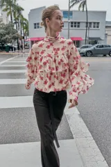 Blusa de cuello subido y manga larga con estampado floral en tonos rosados. Presenta un diseño con volantes en el pecho y a lo largo de las mangas, con cierre posterior mediante botones ocultos.