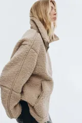 Campera de sherpa suave en color beige, de corte holgado con cuello elevado y cierre frontal bidireccional. Presenta bolsillos frontales tipo canguro y detalles de ribetes en tejido revestido.