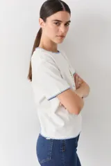 Remera blanca de algodón con cuello redondo y mangas cortas. Presenta un bordado central de un sol amarillo con la palabra 'SAVIA' debajo, y detalles de costura decorativa en color celeste en el cuello, mangas y ruedo.