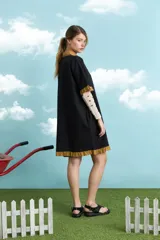 Túnica de algodón color negro, con cuello polo, manga corta, cartera frontal con botones y bolsillo ojal en el pecho. Presenta detalles de voladitos mostaza en el cuello, la botonera, los puños, el bolsillo y el ruedo.