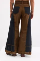 Pantalón de jean estilo wide leg con diseño bicolor en tonos marrón y negro. Presenta paneles laterales oscuros, costuras a la vista en color ocre y bolsillos frontales tipo parche en la parte inferior de las piernas.