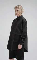 Camisa negra de manga larga, confeccionada en poplín de algodón. Presenta un calce oversize, cuello camisero, cierre frontal de botones y dos bolsillos cuadrados superpuestos en el lado derecho del pecho.