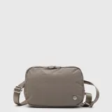 Riñonera Bubba de diseño rectangular, confeccionada en tela resistente color gris topo. Cuenta con cierre superior, correa ajustable para llevar cruzada o a la cintura y detalle de logo metálico en el frente.