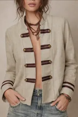 Chaqueta de estilo militar en color beige, con cuello mao y cierre frontal mediante trabas horizontales de color marrón oscuro con botones dorados. Presenta detalles de líneas horizontales en los puños.