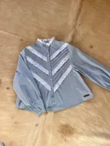Camisa blanca de algodón con cuello alto, mangas largas abullonadas y cierre frontal de botones. Presenta un diseño delantero con alforzas y detalles de bordado inglés en forma de V.