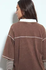 Remera de manga corta color marrón con efecto desgastado, corte oversize y costuras a la vista en color blanco. Presenta cuello redondo con cuello polo blanco superpuesto y mangas largas a rayas horizontales marrones y blancas por debajo.