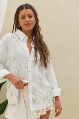 Camisa blanca de manga larga con bordados detallados y lentejuelas plateadas, cuello camisero y cierre frontal de botones.