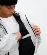 Campera bomber de poliamida en color gris claro, con capucha, cierre frontal con botones a presión y lettering bordado en el pecho. Presenta un diseño funcional con detalles utilitarios.