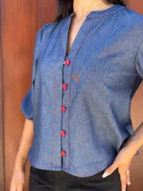 Camisa de jean azul con cuello mao, cierre frontal con botones rojos y mangas tres cuartos abullonadas con puños elásticos.