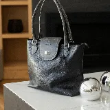 Cartera tote de cuero vacuno color marrón con solapa negra y cierre metálico. Tiene doble asa de mano negra, bolsillo interno con cierre y cierres YKK metálicos.