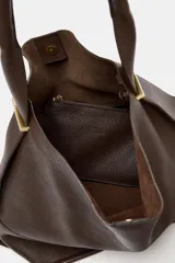 Bolso tipo saco de gran tamaño en cuero marrón oscuro, con textura granulada. Presenta un asa de hombro ancha y detalles metálicos dorados en la unión del asa. Incluye un bolsillo interior extraíble y cierre mediante botón imantado.