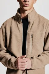Campera deportiva color beige en tejido sherpa, con cuello alto, cierre frontal, bolsillo superior con cierre, bolsillos laterales con cierre, puños y bajo con elástico fino.