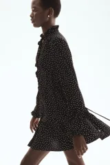 Vestido corto negro con estampado de lunares blancos, escote en V con volados, mangas largas abullonadas y cordón ajustable en la cintura.
