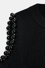 Jersey de punto acanalado color negro, con cuello redondo y mangas largas. Presenta aberturas en los hombros decoradas con detalles de bolas esféricas.