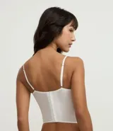 Corset de media copa en color blanco, confeccionado con paneles de poliamida y detalles decorativos de encaje floral en el borde superior y tul en los laterales. Cuenta con tirantes finos ajustables y varillas internas para estructura.