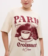 Remera oversize de algodón color beige, de cuello redondo y manga corta, con gran estampado frontal en tonos bordó con temática de París, incluyendo las inscripciones 'PARIS', 'France', 'Croissant & Crème' y un diseño gráfico de una taza de café y un croissant.