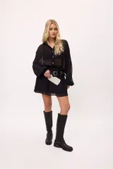 Vestido corto negro de chiffon transparente, con cuello camisero, botones frontales, mangas largas abullonadas y elástico en la cintura. La falda tiene volados.