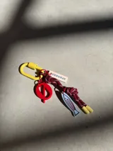 Llavero con mosquetón amarillo, cordón trenzado bordó y dijes colgantes: una placa rectangular color crema con la palabra 'URUGUAY', un dije circular rojo con un símbolo central y un dije en forma de pez color celeste.