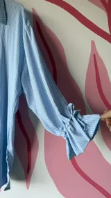 Camisa oversize de fibrana a rayas celestes y blancas, con cuello clásico, mangas largas con puños y botones frontales de acrílico en forma de corazón rojo.