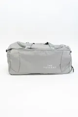 Bolso de viaje gris con doble asa corta y cierre superior. Estampado frontal con la frase "The Tote Bag" en letras blancas.