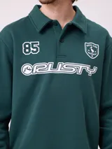 Buzo tipo polo de manga larga color bordó, con cuello camisero y cierre frontal de botones. Presenta estampado gráfico frontal con el número 85, el logo de la marca Rusty y un escudo deportivo.