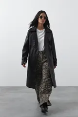 Trench coat de símil cuero color marrón oscuro, con solapas amplias, cierre frontal con botones, cinturón ajustable en la cintura y mangas con tiras ajustables. Presenta un largo por debajo de la rodilla y bolsillos laterales.