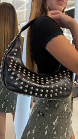 Cartera tipo hobo de cuero sintético negro, adornada con tachas metálicas plateadas en toda su superficie. Cuenta con una correa corta para el hombro y cierre superior.