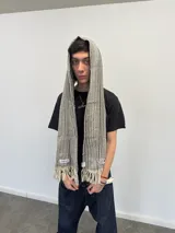 Bufanda larga de tejido trenzado con diseño de cuadros en tonos beige, marrón, negro y blanco. Presenta flecos en los extremos, una grifa de goma con logo en una punta y una etiqueta tejida con logo en la otra.
