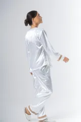 Pijama blanco de satén elastizado, compuesto por una camisa de manga larga con escote en V, abotonada adelante y con puntilla en escote y puños, y un pantalón recto con bolsillos laterales, elástico en la cintura y puntilla en el ruedo.