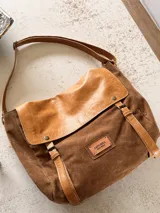 Bandolera tipo messenger bag confeccionada artesanalmente en combinación de cuero y gamuza marrón. Cuenta con solapa frontal de cuero con broches a presión, correas decorativas con hebillas metálicas y una correa larga ajustable para el hombro. Incluye parche con logo grabado en la parte frontal.