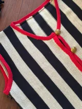 Chaleco de punto a rayas verticales negras y beige, con ribetes rojos en el escote y sisas, cierre frontal con botones dorados grabados.