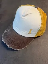 Gorra tipo trucker con visera desgastada en color marrón, panel frontal en color natural y paneles laterales y traseros en color mostaza. Presenta un logo bordado en amarillo en el panel frontal y cuenta con cierre ajustable en la parte posterior.