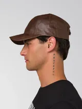 Gorro tipo gorra de béisbol con visera curva, confeccionado en cuero sintético con acabado texturizado. Presenta un pequeño aplique metálico con el logo de la marca en el lateral frontal y cierre metálico ajustable en la parte posterior.